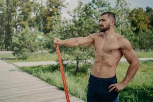 Muscular athletic man has workout with elastic resistance band poses with shirtless muscular body, stands outdoor, concentrated into distance. Athlete sportsman has workout in park. Healthy lifestyle photo