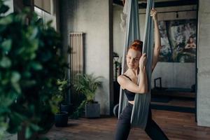 Relaxed red haired woman sitting on aerial yoga hammock and resting after class in fitness studio photo