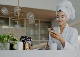 Photo of attractive young woman wears bath towel on head and soft white dressing gown holds mobile phone enjoys online communication applies moisturising undereye patches drinks hot beverage