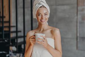 Attractive smiling young woman with makeup, toothy smile, poses bare shoulders, wrapped in towel, drinks hot beverage stands indoor has cheerful expression applies moisturizing cream on face photo