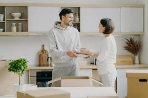 Positive young husband and wife unpack personal belongings in kitchen, carry plates, look gladfully at each other, poses indoor against cozy interior, happy to move into new modern flat or house photo