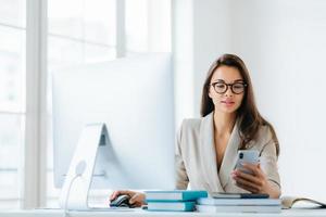 Elegant woman office worker uses mobile phone and computer at one time, sends messages and chats with clients online, monitors news from networks, sits at desktop with pile of notepads and books photo