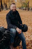 Vertical image of smiling motorcyclist smiles happily, holds helmet, poses on motorbike, wears black jacket and jeans, has drive through park, enjoys autumn time rides black vehicle. Transport concept photo