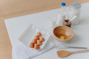 Cooking ingredients on kitchen table. View from above of eggs, milk and flour, whisk and wooden spatula near. Utensils and fresh products photo