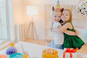 Friendly two girls embrace and have good relationship, stand near festive table with cake, celebrate birthday together, stand in living room. Glad female sisters enjoy holiday, special occasion photo