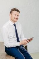 Confident pleased young male entrepreneur in luxury formal clothes, reads recieved text message from colleague on modern tablet computer, looks joyfully at camera, poses in office. Vertical shot photo