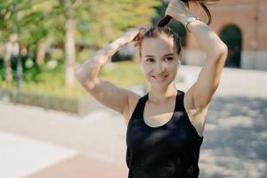 Positive sportswoman combes pony tail being in good physical shape listens music in earphones wears casual t shirt looks somewhere poses outdoor enjoys good weather sunny day. Sport concept. photo