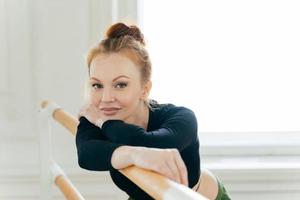 Headshot of calm beautiful female ballet dancer with ginger bun, wears sportswear, has flexible body, poses near bars, develops dancing skills, has minimal makeup. Professional ballerina in studio photo