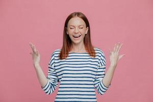 Excited overjoyed woman closes eyes, keeps hands raised, dressed in striped sweater, laughs positively, isolated over pink background. People, happiness, joy concept. Glad young female model photo