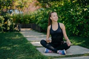 Dark haired young European woman sits crosses legs looks aside with satisfied expression uses smartphone has break after cardio training dressed in active wear and sneaker reads text message photo