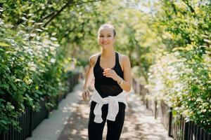 una joven deportista tiene un entrenamiento para trotar corre al aire libre sonríe agradablemente disfruta de un buen día vestida con ropa deportiva escucha música de la lista de reproducción. Comience el día desde el trote matutino. corredora foto