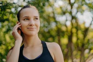 Healthy lifestyle concept. Outdoor shot of sportswoman enjoys favorite playlist in earbuds while training in park has gentle smile concentrated into distance poses over blurred nature background photo