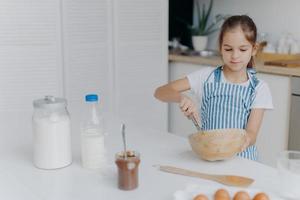 Children, cooking and home concept. Pretty dark haired girl whisks ingredients in bowl, busy preparing dough for cake, being future chef, wears white t shirt and striped apron, uses ingredients photo