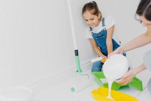 Happy mother and small daughter make repairing at their home, going to paint walls, pour white paint in tray, poses in spacious room, busy with house renovation, redecoration or refurbishment photo