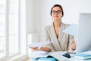 Busy successful female manager works with papers in office, poses at desktop, wears spectacles and formal outfit, busy preparing report, looks confidently at camera. Businesswoman reviews documents photo