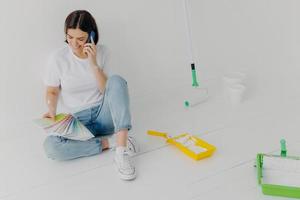 Satisfied female designer discusses ideas via smartphone, looks attentively at color samples, sits on floor in empty room which needs redecoration, has cheerful mood, chooses colour for painting photo