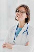 Close up portrait of short haired female general practitioner stands with smile and arms crossed, uses stethoscope, enjoys work, poses against white background. People, mediccare and treatment concept photo