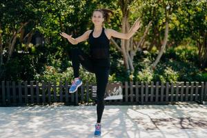 Outdoor shot of athletic motivated woman jumps high does physical exercises keeps hands and legs raised dressed in black sport clothes being in good mood being full of energy. Sport lifestyle photo