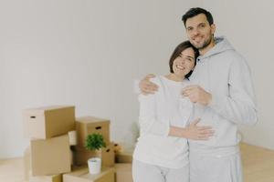 Couple in love purchase own house, embrace and stand closely to each other, hold keys from new apartment, carton boxes in background, have delighted face expressions. Satisfied clients move in photo