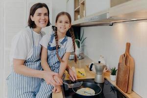 madre complacida con una pequeña hija que ayuda cerca de la cocina, preparar el desayuno para toda la familia, freír huevos en la sartén, posar en la cocina moderna. niño pequeño aprende a cocinar de mamá. concepto de familia y comida foto