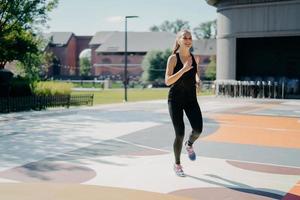 Cheerful athletic woman runs actively on stadium dressed in black sportclothes enjoys physical activities outdoors during summer day being full of energy. People sport and motivation concept photo