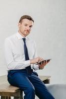Serious male entrepreneur watches video on digital tablet, messages with business partners, dressed elegantly, sits on wooden table, poses against white wall. Businessman updates banking account