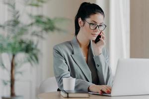 Serious brunette woman keyboards information, concentrated into laptop computer, dressed in formal outfit, poses at coworking space, works remotely, involved in working process during daytime. photo