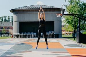 Athletic young woman in black activewear jumps high keeps arms raised up clasps hands warms up before jogging has training on stadium outdoors has cardio burns fat and calories for weight loss photo