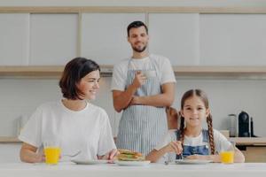 un niño pequeño encantador y su madre desayunan juntos, se sientan en la mesa de la cocina, comen una comida deliciosa, el padre se para en el fondo, usa delantal y toma café. la familia amistosa se reúne en la cocina foto