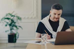 Male office worker looking at laptop and making notes while sitting in office photo