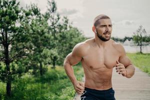 Sporty athletic man runs outdoor with naked torso, leads healthy lifestyle, has morning workout, listens music in wireless earphones, has muscular body, breathes fresh air. Health activities photo