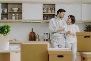 Indoor shot of lovely newlywed couple talk to each other with pleased expressions, drink takeaway coffee from paper cups, pose in modern kitchen with new bought furniture, change place of living photo