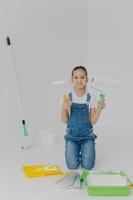 Happy little girl in denim overalls stands on knees, holds paint roller, surrounded with trays with paint, poses in white room, has happy expression, helps parents with house improvement and painting photo