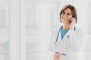 Shot of pleased female physician or surgeon gives prescription via mobile phone, has conversation with patient, holds pen, wears white uniform, works in hospital, stands indoor against window photo