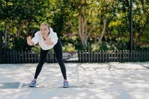 Outdoor shot of satisfied healthy sporty woman leans forward keeps palms raised does exercises in open air trains in park feels good wears sweatshirt leggings and sneakers leads active lifestyle photo