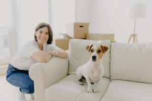 Happy woman plays with favourite pet, poses near sofa in new apartment, celebrate Moving Day, bought new house, pose in spacious white living room, settling furniture, think about interior design