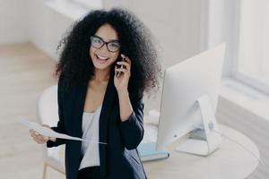 Cheerful female business worker calls partner to discuss details of statrup project, holds paper documents, wears optical glasses and elegant suit, gets necessary information, dressed elegantly photo