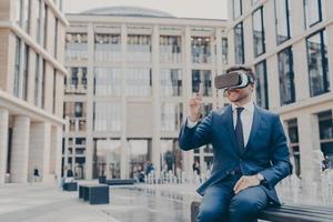 Smiling businessman with stubble sitting on bench in town square with VR glasses on photo