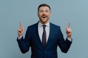 Positive optimistic businessman points above demonstrates something with glad expression dressed in formal suit white shirt and tie poses against blue background shows top advertisement deal on market photo