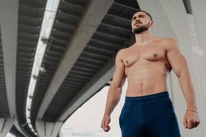 View from bottom of self confident bearded handsome European man has strong perfect body looks into distance, leads active lifestyle, poses outside under bridge. Young shirtless bodybuilder. photo