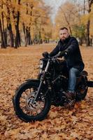 Brutal male biker rides motorcycle during autumn time, poses in park with many trees and yellow foliage, enjoys driving in open air, has tour journey. Motorcyclist with own transport outdoor photo