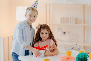 Happy small girl and boy hold gift box with red ribbon, prepare surprise on birthday, pose at white table, wear cone party hats, have positive expressions. Childhood concept photo