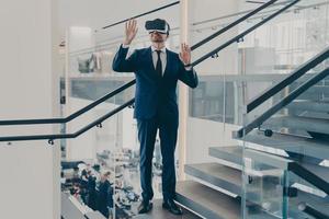 Businessman in VR headset stands on staircase in office lobby with hands up in air photo