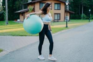 Full length shot of sporty slim woman holds big inflated fit ball, dressed in active wear, has aerobics exercises outdoors, has athletic body stands on road with house in background. People, lifestyle photo