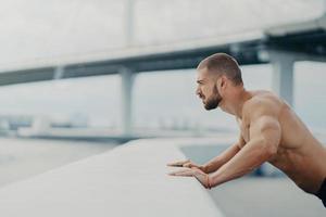 Sideways shot of handsome muscular man with thick beard does push ups exercise has workout outdoor leans on bridge fence engaged in physical activity poses with bare torso has strong muscles photo