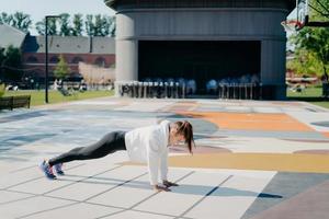 Full length side view shot of young active woman stands in plank position dressed in sweatshirt leggings sneakers has training outdoors on stadium goes in for sport. Morning workout concept. photo