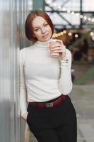 Vertical image of thoughtful young woman wears white jumper and black trousers, holds takeaway coffee, dreams about something pleasant, poses outdoor, keeps hand in pocket. People and lifestyle photo