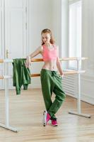 Beautiful fit ginger female dancer has rest near barre in ballet dancing studio, focused down with thoughtful expression, wears top, green trousers and sneakers, prepares for performance on stage photo
