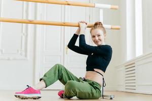 Female gymnast sits on floor, dressed in sport outware, stretches herself near barre, holds hand on handrail, looks with satisfied expression at camera, shows fit body. Dancing, flexibilty, aerobics photo