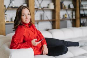 Photo of attractive female model in knitted red sweater, messages online at home, poses on couch in living room, uses modern cell phone and wireless internet connection, looks with appealing look
